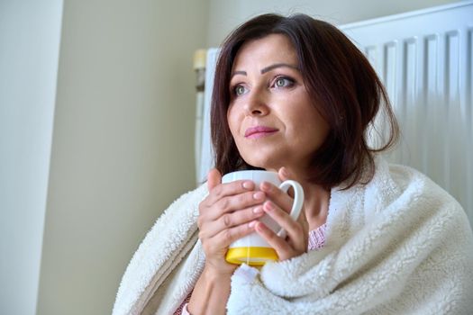 Cold winter autumn season at home, frozen woman basking herself with a warm blanket and a mug of tea near the central heating radiator