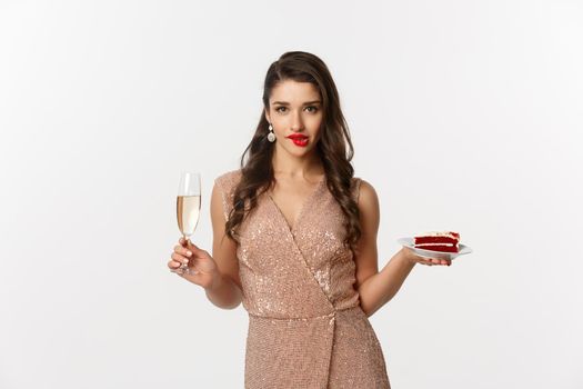Party and celebration concept. Sexy woman in elegant dress, holding champagne and piece of cake, biting lip and looking seductive at camera.