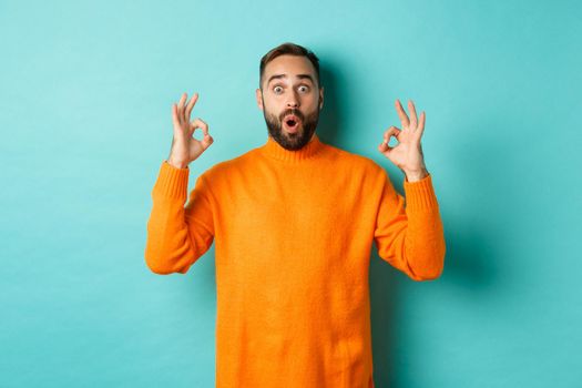 Impressed man praising something super cool, showing okay sign and looking wondered at camera, light blue background.