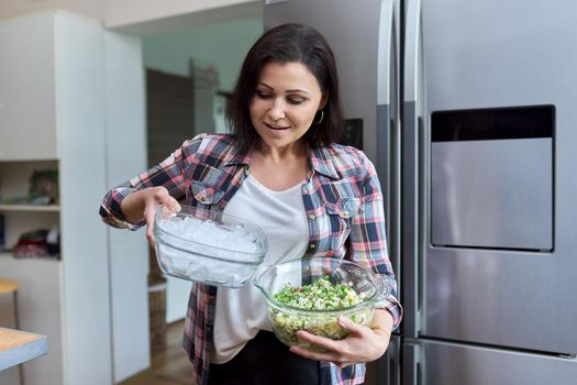 Middle aged woman in the kitchen with salad and a bowl of ice in the kitchen near the refrigerator. Homemade food, lifestyle, home, people concept