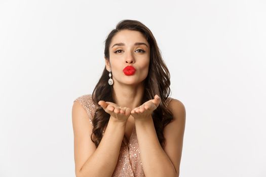 Concept of New Year celebration and winter holidays. Close-up of sensual young woman in dress, pucker lips and blowing air kiss at camera, white background.