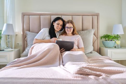 Mother and teenage daughter resting at home together, use digital tablet. Woman and teen girl lying in bed, looking at screen, movie video. Family, parent teenager relationship, vacation, technology