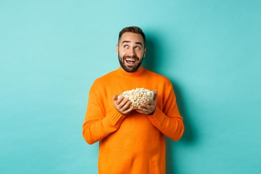 Handsome young man in orange sweater, looking thoughtful at upper left corner, holding popcorn, picking movie, blue background.