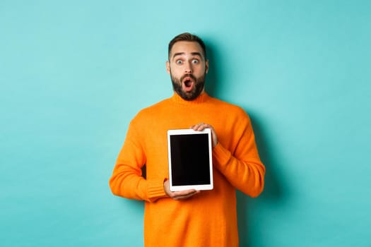 Technology. Surprised bearded man drop jaw, staring at camera and showing digital tablet screen, standing againt light blue background.