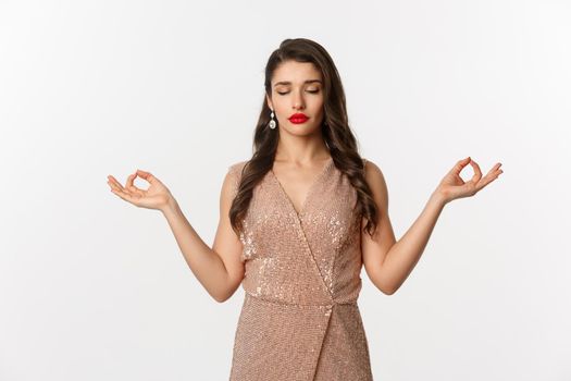 Christmas party and celebration concept. Calm and patient young woman in stylish dress meditating, feeling zen, standing over white background.