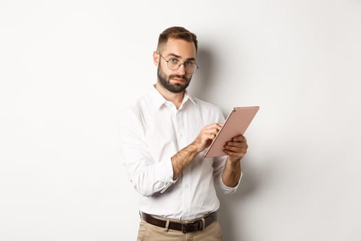 Handsome businessman doing job on digital tablet, reading something, standing over white background.