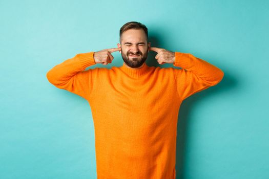 Displeased man shut ears and grimacing, loud neighbours, standing annoyed with noise, light blue background.