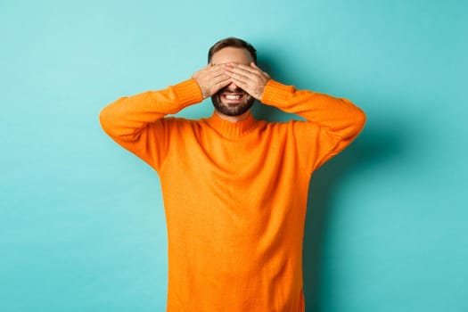 Happy bearded man waiting for surprise, shut ears and expeting gifts, standing over light blue background.