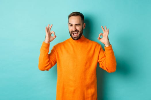 Confident man assuring all ok, showing okay signs and winking at you, recommending product, standing in orange sweater at studio background.