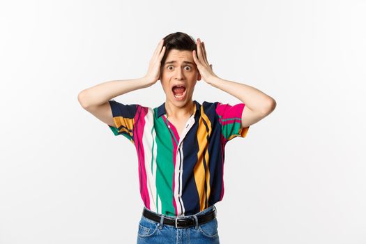 Scared young man screaming in panic, looking alarmed and holding hansd on head, standing troubled and worried against white background.