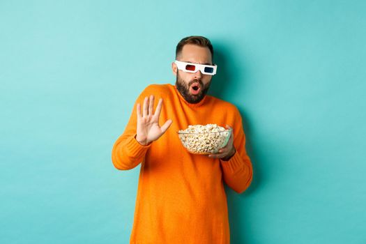 Image of man in 3d glasses watching movie, getting scared of special effects, looking in awe, standing with popcorn against white background.