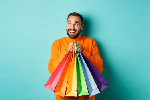 Happy caucasian man in orange sweater, looking left and imaging, holding shopping bags and smiling, standing over turquoise background.