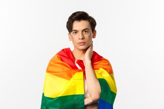 Waist-up shot of beautiful gay man with glitter on face, wrap himself with rainbow lgbt flag and touching face gently, looking at camera, white background.