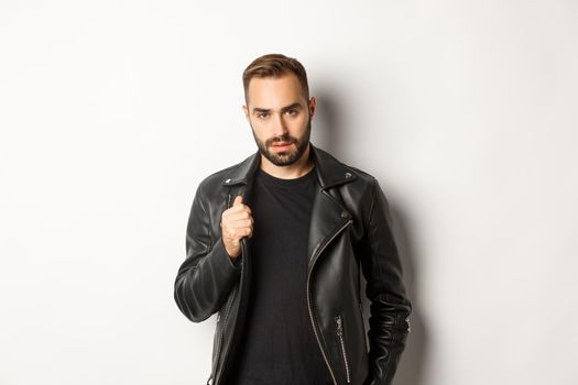 Handsome bearded man expressing confidence, touching his leather jacket and looking self-assured, white background.