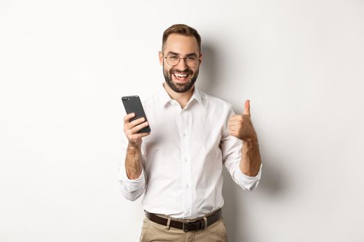 Satisfied business man showing thumbs up after using mobile phone, standing pleased over white background.