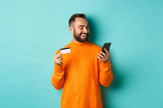 Online shopping. Handsome bearded man paying in internet, holding credit card and stare at mobile screen, standing over turquoise background.