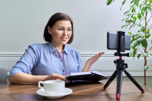 Middle aged business woman, counselor, mentor in video meeting, using smartphone on tripod. Positive female with cup of coffee talking looking in webcam. Video consultations, freelancing, remote business, online education