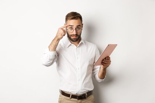 Displeased employer scolding staff while checking report on digital tablet, pointing at head and looking disappointed, standing over white background.