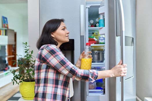 Woman opens refrigerator at home in kitchen, holding bottle of orange juice. People, food, drinks, lifestyle, home, kitchen, refrigerator, groceries concept