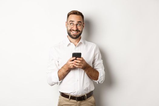 Thouthful handsome manager using mobile phone and thinking about answering message, looking at upper left corner and smiling, standing over white background.