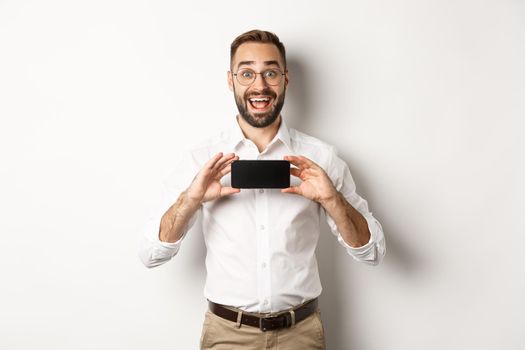 Excited handsome man showing mobile phone screen, looking amazed, standing over white background.