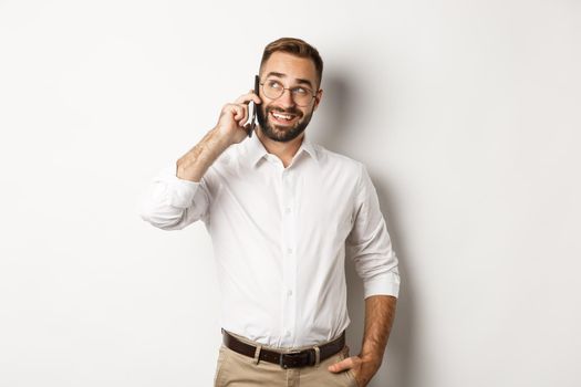 Successful business man in glasses talking on mobile phone, looking satisfied and smiling, standing against white background.