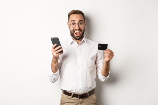 Business and online payment. Excited man paying with mobile phone and credit card, smiling amazed, standing over white background.