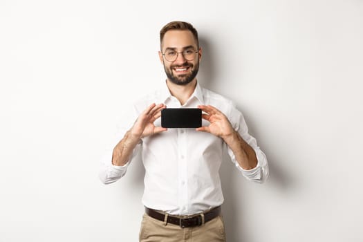 Handsome bearded man in glasses, demonstrating mobile phone application, showing smartphone screen, standing over white background.