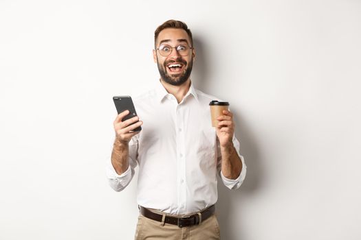 Amazed businessman drinking coffee, reacting at awesome online offer on mobile phone, standing over white background.