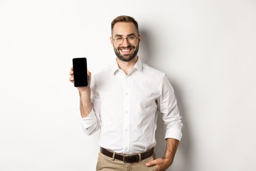 Satisfied business man showing mobile screen, smiling proudly, standing over white background.