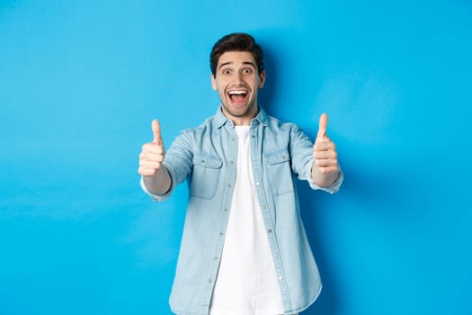 Smiling confident man showing thumbs up with excited face, like something awesome, approving product, standing against blue background.