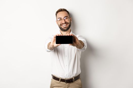 Happy business man showing mobile screen, holding phone horizontally, standing satisfied against white background.