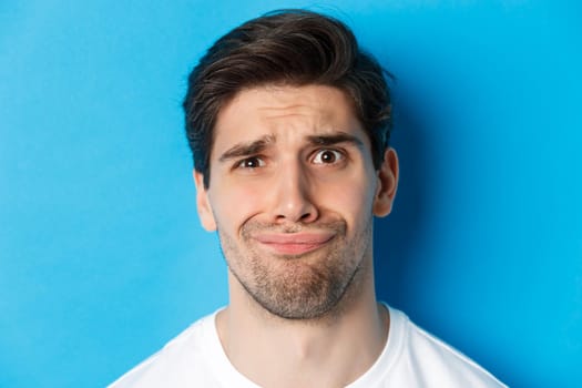 Head shot of skeptical and disappointed man, grimacing complicated, standing against blue background.