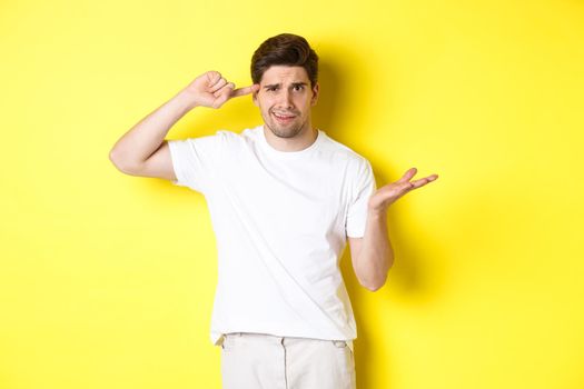 Confused and angry man pointing at head, scolding person for acting stupid, show crazy sign, standing over yellow background.