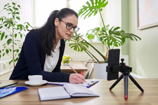 Middle aged business woman, teacher working from home remotely, using smartphone for video call, video conference. Freelance, distance business, technology, home Office, people concept