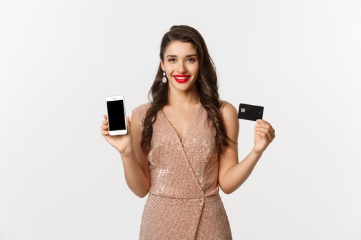 Online shopping and holidays concept. Attractive woman in party dress showing credit card and mobile screen, smiling happy, white background.