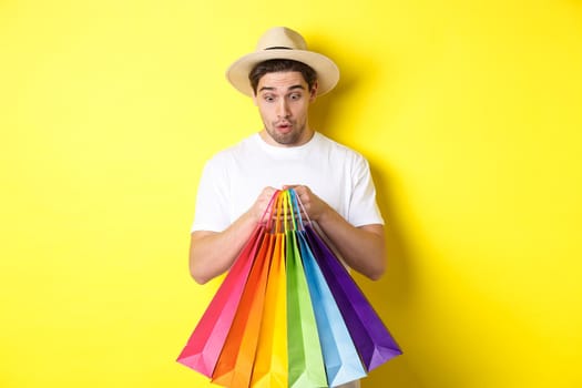 Happy man looking surprised at shopping bags, buying souvenirs on vacation, standing over yellow background.