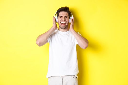 Happy guy listening music in new headphones, buying earphones on black friday, standing over yellow background.