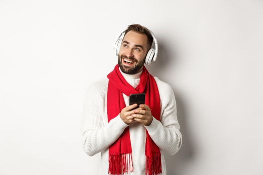 Winter holidays and technology concept. Happy man listening music podcast on headphones, holding mobile phone and looking at empty space, white background.