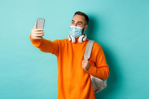 Young modern man with headphones and backpack, taking selfie on mobile phone in medical mask, standing over light blue background.