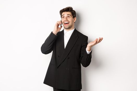 Portrait of happy good-looking businessman receiving great offer, talking on phone and looking pleased, standing in black suit against white background.