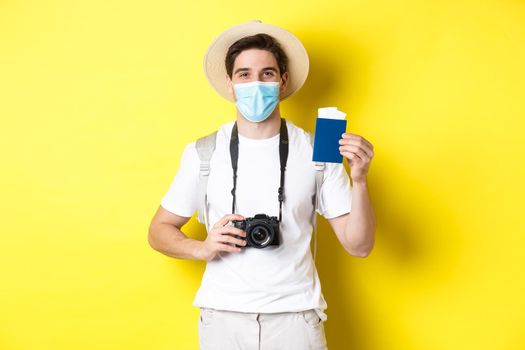 Concept of covid-19, travelling and quarantine. Happy man tourist with camera, showing passport and tickets for vacation, going on trip during pandemic, yellow background.