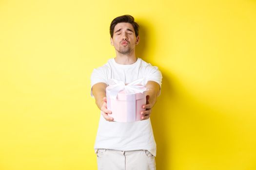 Concept of holidays and celebration. Boyfriend giving a gift, close eyes and pucker lips for kiss, standing over yellow background.