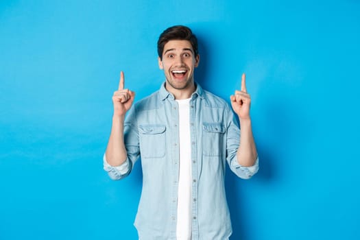 Portrait of happy 25s guy with beard, pointing fingers up and smiling, showing advertisement, standing against blue background.