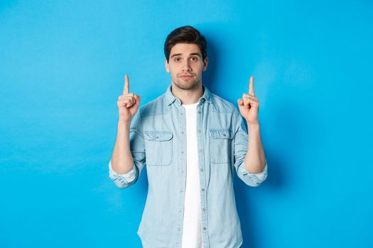 Image of calm handsome man showing you promo offer, pointing fingers up at copy space, standing against blue background.