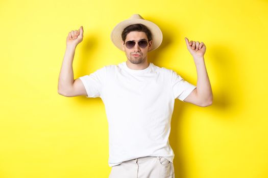 Tourism, travelling and holidays concept. Man tourist enjoying vacation, dancing in straw hat and sunglasses, posing against yellow background.