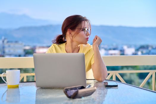 Serious middle aged woman with laptop at table on outdoor balcony. Concentrated 40s mature business woman working remotely. Freelance, technology, work from home office, online training, blog, vlog