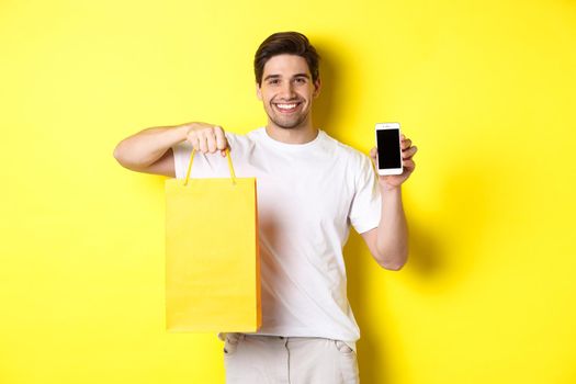 Concept of mobile banking and cashback. Young happy guy holding shopping bag and showing smartphone screen, yellow background.