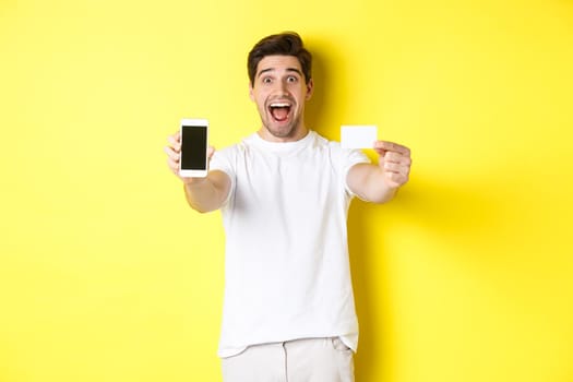 Handsome caucasian man showing smartphone screen and credit card, concept of mobile banking and online shopping, yellow background.