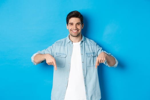 Handsome smiling adult man introduce product, pointing fingers down at promotion, standing against blue background.